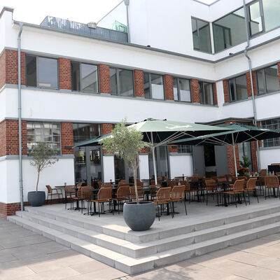 Bild vergr&ouml;&szlig;ern: Blick auf ein weiß gestrichenes Gebäude, dessen Fenster mit roten Klinkersteinen eingefasst sind. Vor dem Gebäude liegt eine Terrasse, auf der Tische und Stühle unter Sonnenschirmen stehen.