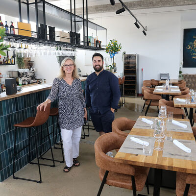 Bild vergr&ouml;&szlig;ern: Blick in den Gastraum eines Restaurants, links eine lange Theke, die mit blauen Kacheln verkleidet ist, in der Mitte stehen eine Frau und ein Mann nebeneinander und schauen in die Kamera, rechts sind hölzerne Tische und Stühle zu sehen.