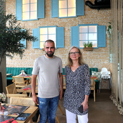 Bild vergr&ouml;&szlig;ern: Ein Mann und eine Frau stehen nebeneinander in einem Raum neben einem Tisch, im Hintergrund sind ein Olivenbaum sowie eine gemauerte Wand und darin beleuchtete Fenster mit aufgeklappten, hölzernen Fensterläden zu sehen.