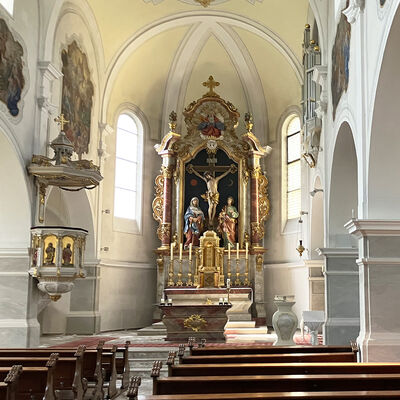 Bild vergr&ouml;&szlig;ern: Blick in den Innenraum einer Kirche, im Vordergrund hölzerne Bänke, hinten der Altar mit goldumrahmtem großem Altarbild, das die Kreuzigung Jesu zeigt. Durch die großen Kirchenfenster fällt Sonnenlicht.