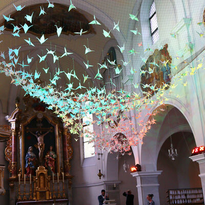 Bild vergr&ouml;&szlig;ern: Gezeigt wird der Innenraum einer Kirche. Unter der Decke ist ein Netz gespannt, in dem zahlreiche aus Papiergefaltete Vögel angebracht sind. Diese werden in verschiedenen Farben angeleuchtet.