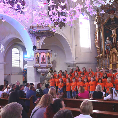 Bild vergr&ouml;&szlig;ern: Das Bild zeigt Menschen, die in einer Kirche sitzen und einem Kinderchor lauschen, der vorne im Altarbereich steht. Die Kinder tragen alle orangefarbene T-Shirts. An der Decke hängt ein Netz, in dem aus Papier gefaltete Vögel angebracht sind.